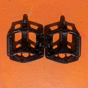 A pair of metal bicycle pedals in a sleek, modern design, featuring a silver finish and textured surface for grip, displayed on a plain background.