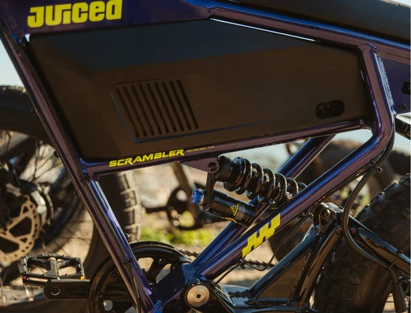Electric bike parked outdoors, featuring a retro-style frame, fat tires, and a long padded seat, with "Juiced Scrambler" branding visible on the battery.