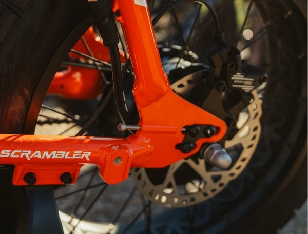 Electric bike parked outdoors, featuring a retro-style frame, fat tires, and a long padded seat, with "Juiced Scrambler" branding visible on the battery.
